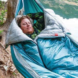 Person sleeping in a blue and gray sleeping bag in a hammock with a mountainous background