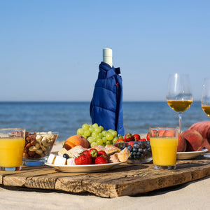 Outdoor picnic setup with fruits, juice, and wine on a wooden board by the sea.