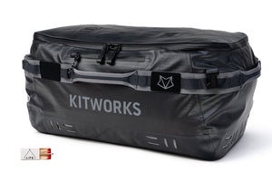 A dark gray waterproof duffel bag with 'KITWORKS' printed in white on the front, featuring a top zipper, side straps, and a small logo patch. A small inset graphic in the bottom-left corner shows a tent and a red-and-white striped item.