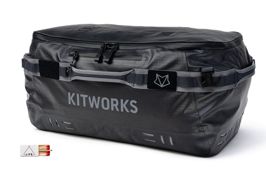 A dark gray waterproof duffel bag with 'KITWORKS' printed in white on the front, featuring a top zipper, side straps, and a small logo patch. A small inset graphic in the bottom-left corner shows a tent and a red-and-white striped item.